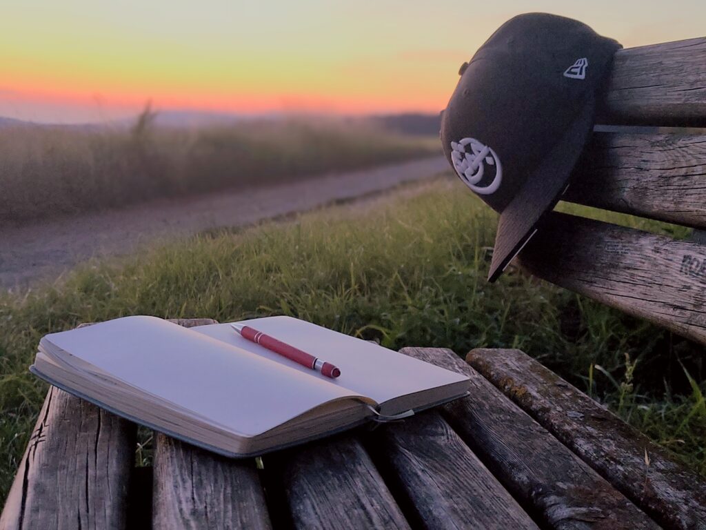 Aufgeschlagenes Notizbuch mit rotem Stift auf einer Holzbank im Abendlicht, daneben eine schwarze Cap mit AZAD Logo.