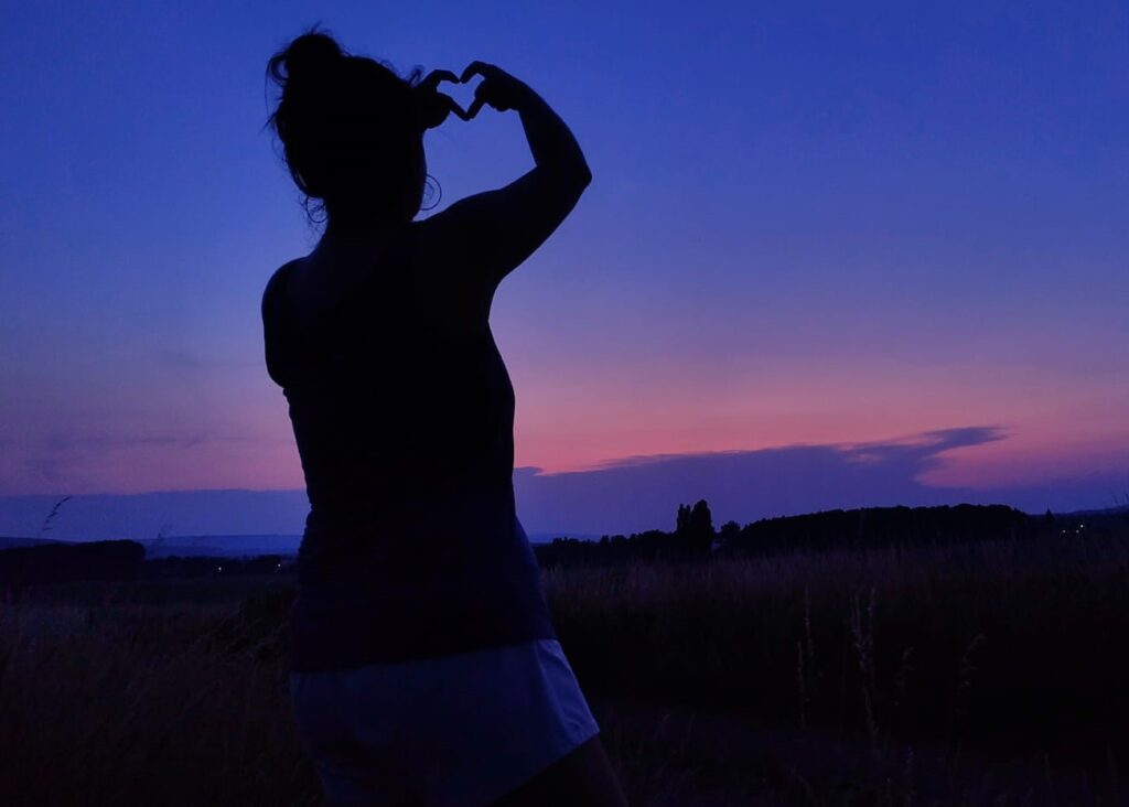 Silhouette mit Herzgeste vor blauviolettem Abendhimmel mit rosa Streifen.